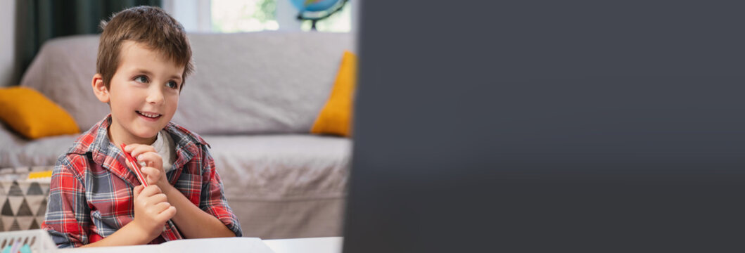 A preschool boy learns to write capital and cursive letters in a copybook at home, sitting at a desk near a laptop. Online tutor, distance learning banner copyspace