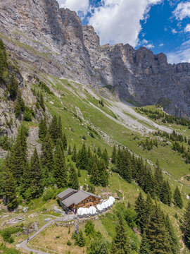 Aerial view of Tallihutte SAC mountain hut nestled on a green alpine slope with pine trees and a dramatic rocky mountain face in Innertkirchen, Bern, Switzerland.