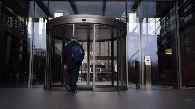 Man with backpack walking away from camera and entering modern office building through revolving door, view from interior lobby, professional and contemporary business concept