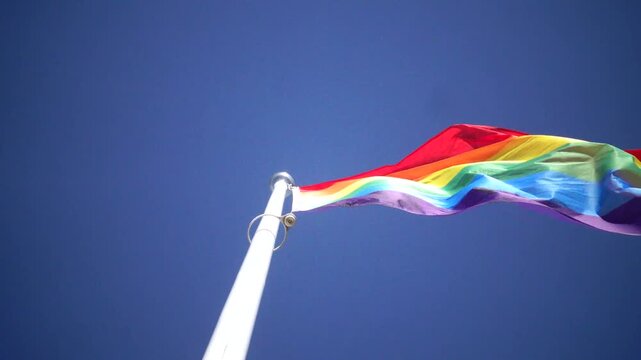 Stockholm, Sweden A Pride flag flies on a flagpole against a blue sky. 