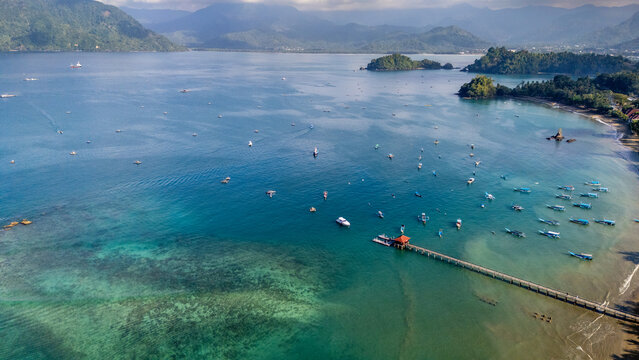 Aerial view of Pantai Karanggongso, featuring a long wooden pier in the clear turquoise bay, surrounded by small fishing boats and lush mountains in Watulimo, East Java, Indonesia.