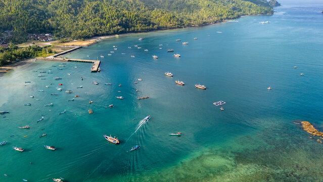 Aerial view of Pantai Karanggongso featuring numerous small fishing boats scattered across the turquoise bay, a wooden pier, and lush green hills in Watulimo, East Java, Indonesia.