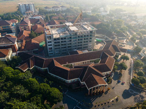 Aerial view of a building complex under construction with a concrete frame surrounded by buildings with brown tiled roofs and trees in Banyumas Regency, Central Java, Indonesia.