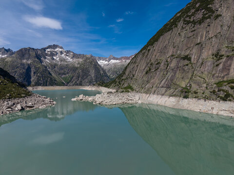 Aerial view of Gelmersee reservoir in the Swiss Alps, showing low water levels, rocky shoreline, and surrounding mountain peaks under a clear blue sky in Switzerland.