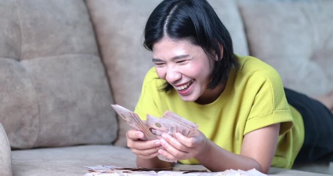 Happy Asian woman counting cash money lying on sofa at home, Thai baht banknote, money, cash, financial success, savings, income, bonus, wealth, and economic stability with cheerful emotion