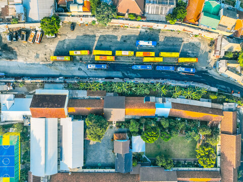Aerial view of Baranangsiang bus terminal with rows of yellow and blue buses parked in the lot, surrounded by lush trees and urban buildings in Bogor Timur, West Java, Indonesia.