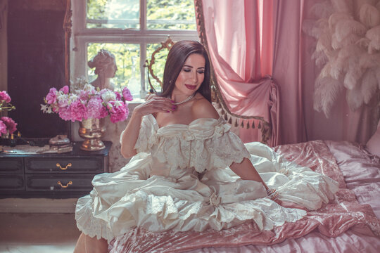 Sensual dark-haired lady posing on a velvet bed surrounded by blooming pink peonies, luxury fragrance and beauty campaign concept in a vintage boudoir setting