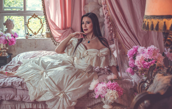 Sensual dark-haired lady posing on a velvet bed surrounded by blooming pink peonies, luxury fragrance and beauty campaign concept in a vintage boudoir setting