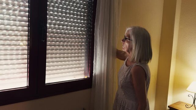 An elderly woman raising a window blind early in the morning to let in the daylight.
