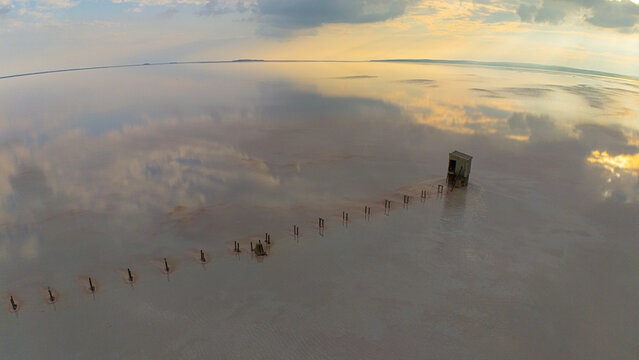 Aerial view of Koyuncu Salt Works at Lake Tuz with a small wooden structure and a line of posts extending into the pink salt water under a sunset sky in Sereflikochisar, Ankara, Turkiye.