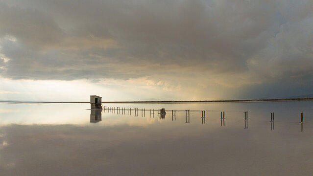 Aerial view of Koyuncu Salt Facility with a small structure and wooden posts in the calm, reflective waters of the salt lake under a dramatic cloudy sky in Sereflikochisar, Ankara, Turkiye.