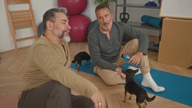 Man high fives his partner over a blue yoga mat while holding a water bottle and greeting a small dog in a studio; friendship relaxation.