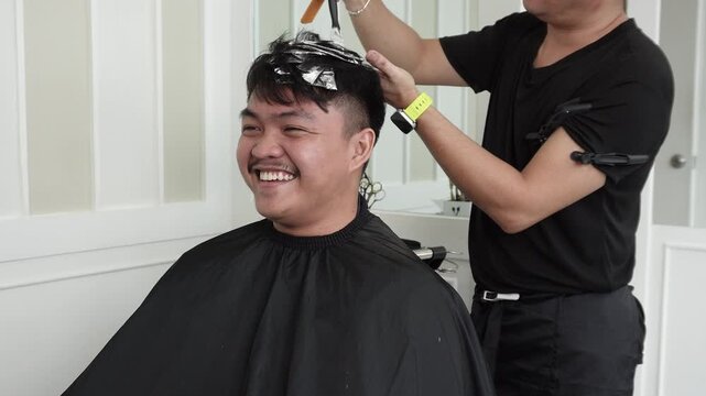 Hair stylist applying lightener to foil sections during professional hair highlights treatment for Southeast Asian man in his early 20s