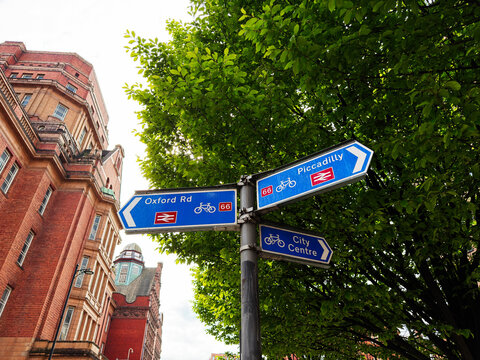 Blue cycle signpost pointing to Oxford Rd, Piccadilly and City Centre beside red brick building and trees in Manchester, UK