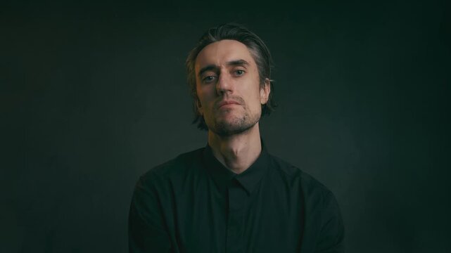 Young adult man with medium-length hair in black collared shirt posing for moody studio headshot against dark gradient background