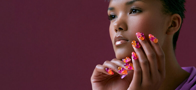 Woman with colorful gummy candy manicure posing against burgundy background, playful beauty trend with vibrant nail art and stylish makeup