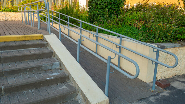 Side view of paved wheelchair ramp featuring light blue painted metal handrails installed next to stone steps leading to public facility entrance