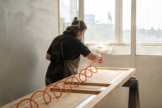 Professional carpenter blows off dust from restored antique wooden door with air hose in paint booth, craftswoman in safety gear using spiral hose for supplying air to remove dirt before painting.