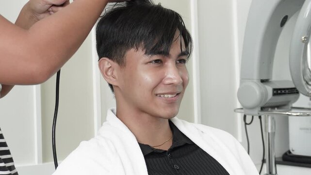 Hair stylist blow drying and round brushing hair after professional cellophane treatment for Southeast Asian man in his early 20s