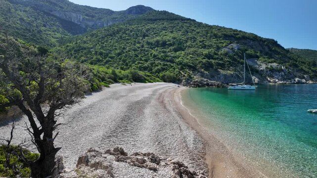 Sveti Ivan beach, pebble cove on Cres Island, Croatia, showing turquoise crystal clear Adriatic sea water, surrounded by green hills, with a sailboat anchored near coastline in Cres Island of Croatia