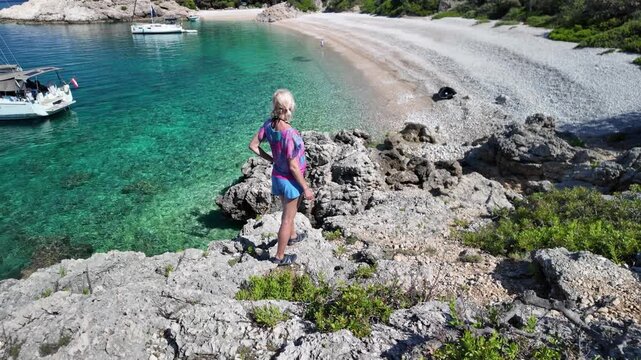 Woman relaxing on a rocky coast, overlooking Sveti Ivan Beach with its crystal clear Adriatic Sea and moored sailboats, enjoying a peaceful summer day in Lubenice, Cres Island of Croatia