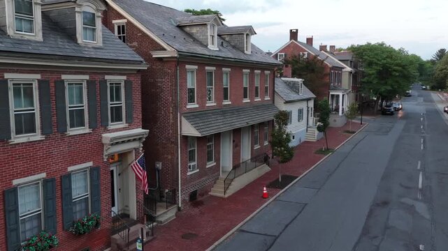 American flag hangs beside brick rowhouse entrance on historic tree-lined street in Bethlehem Pennsylvania, dormer windows and slate roofs line brick paved sidewalk below. Aerial drone view.