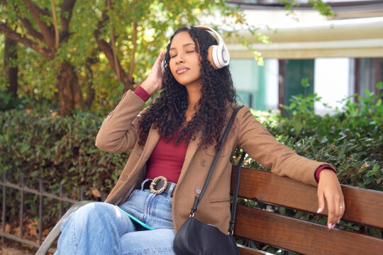 Young lady relaxes outdoors. Calm woman immersed in music on park bench with greenery
