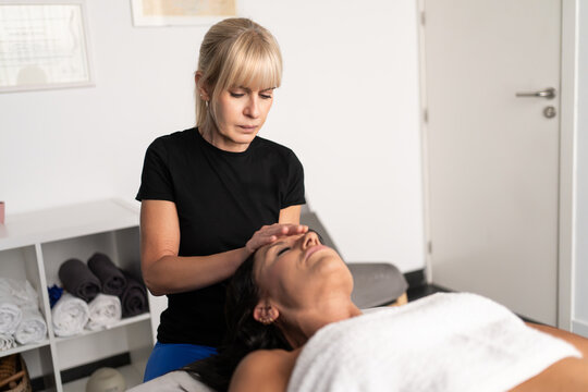 Female therapist performing gentle facial osteopathic treatment for relaxed adult woman lying on treatment table inside bright minimal clinic room, focused hands supporting wellness care