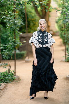 Fashionable woman in flowing attire stands confidently on treefringed route during twilight