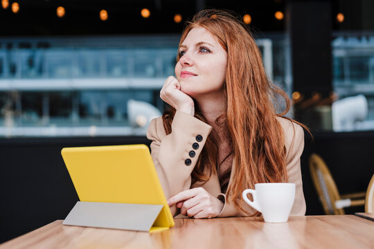 Beautiful woman with hand on chin day dreaming while sitting in front of digital tablet and coffee cup at cafe