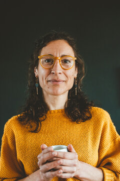 Smiling mature woman wearing eyeglasses holding coffee cup against black background