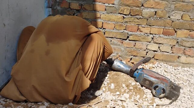 Construction worker using jackhammer on ground, laborer breaking concrete with pneumatic drill, manual labor at building site with brick wall background