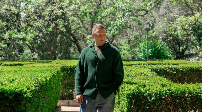A man in a green sweater stands in a park lost in thought