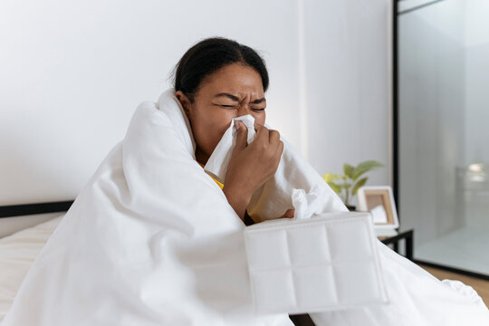 Sick woman sitting on bed wrapped in a blanket blowing nose in a tissue