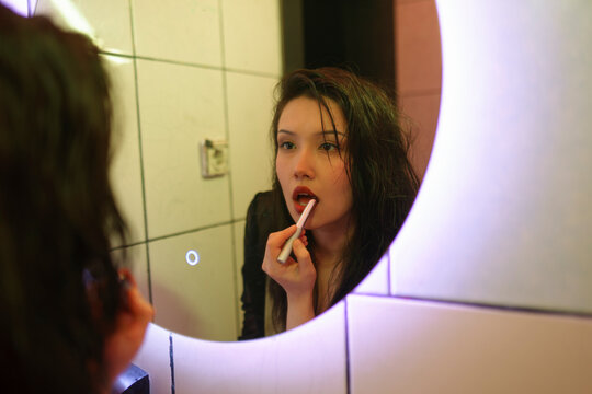 Woman applying red lipstick in illuminated mirror at evening