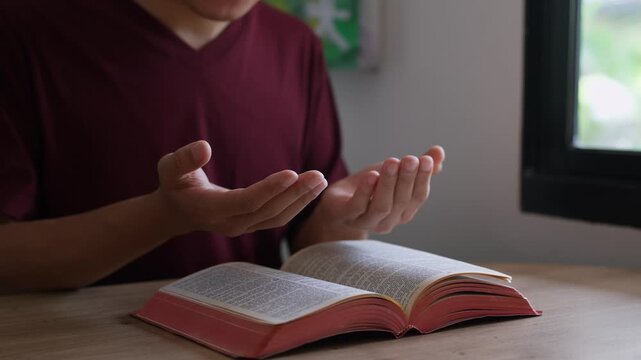 A Christian man raising open hands in praise and prayer over Holy Bible on a table indoors. Spiritual surrender, Pray to God to confess your sins and ask for blessings to overcome his problems. 