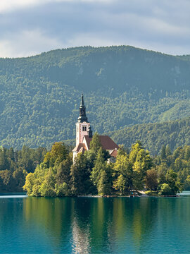 Scenic view of Lake Bled with the iconic island church reflected in turquoise water, surrounded by lush forests and Alpine mountains on a sunny day in Slovenia
