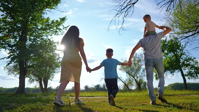 Happy family walks together holding hands in park. Father carries child on shoulders. Parents and son stroll along grassy path among trees. Family bonding walk in sunny nature at summer sunset.