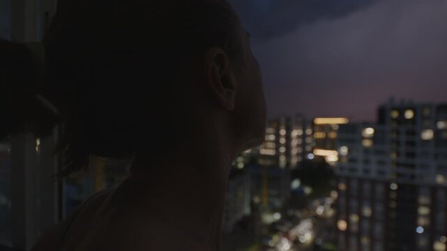 Storm over the city. Woman watches thunder storm with lightnings from the balcony of her apartment on a top floor with city view on the background