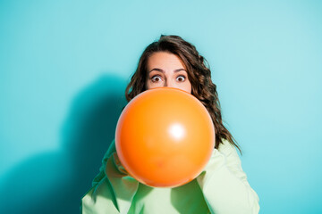 Obraz premium Young woman blowing up a large orange balloon with surprised expression against a turquoise background for playful party and celebration concept