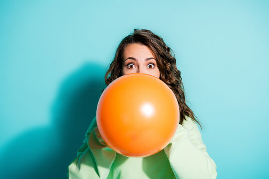 Young woman blowing up a large orange balloon with surprised expression against a turquoise background for playful party and celebration concept