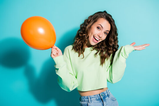 Happy young woman holding an orange balloon and smiling against a turquoise background, casual fashion portrait conveying joy and celebration