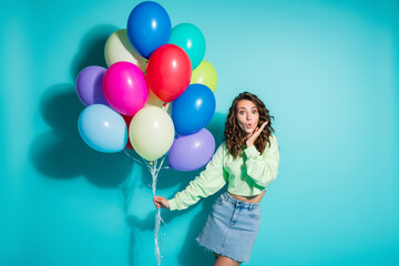 Obraz premium Young woman holding colorful balloons and looking surprised against a turquoise background in a casual denim skirt at a cheerful party celebration