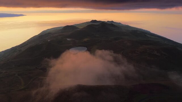 Aerial view of the volcanic landscape of Pico Island with a crater lake partially covered by clouds at sunset in Pico, Azores, Portugal.