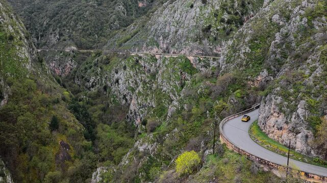 Aerial view of a yellow car driving along a winding mountain road through a deep rocky canyon with lush green vegetation in Anversa degli Abruzzi, Abruzzo, Italy.