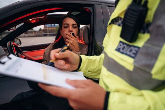 Female driver in car smoking while police officer in reflective jacket writes ticket on clipboard during traffic stop in urban environment