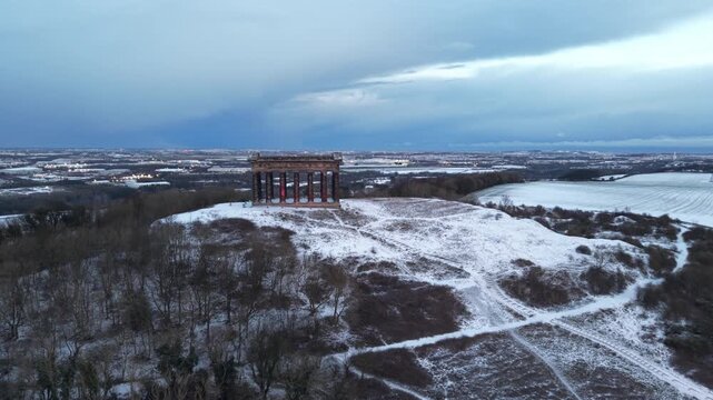 Aerial drone shot pushing slowly in toward the snow-covered Penshaw Monument in Sunderland, with the Nissan factory visible across the winter landscape in the background.