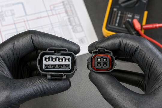 Mechanic holding electric car connector in gloved hand. Auto electrician repairing vehicle wiring system. Diagnostic maintenance of automotive component with multimeter and circuit diagram.