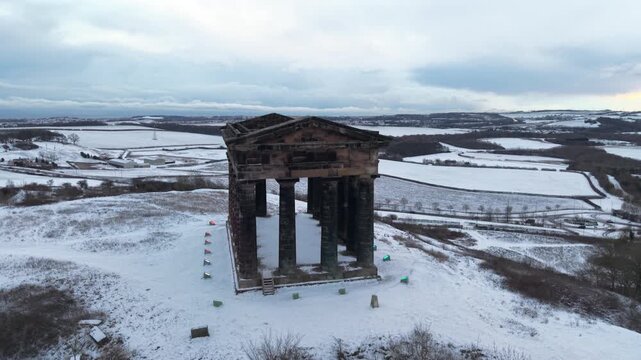 High-angle aerial drone shot rising over and tracking along the Penshaw Monument on a snow-covered hill in Sunderland during a cold winter day.