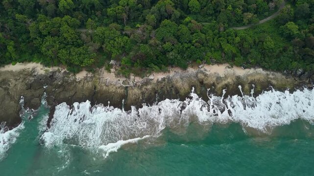 Aerial view of waves crashing against the rocky coastline bordered by a dense tropical forest in Indonesia.